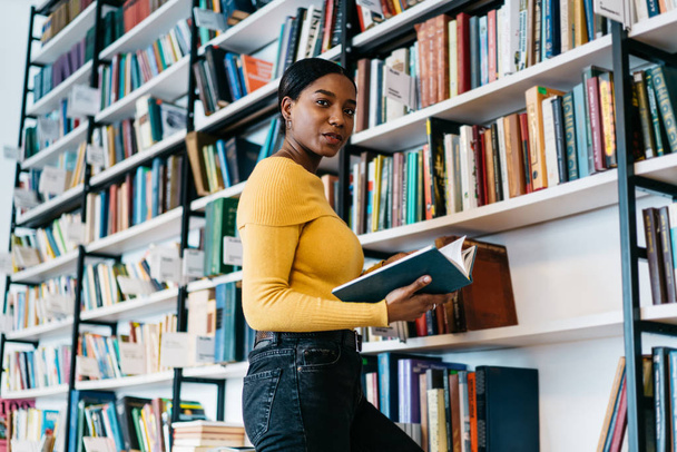 Portrait of positive african american young woman holding literature book in hands and smiling at camera.Dark skinned student preparing for writing course work searching information in textbook - Фото, зображення