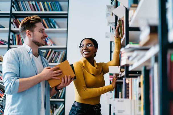 Positive two multicultural young people teamworking in library.Cheerful diverse hipster students dressed in formal wear pulling books from shelf choosing literature for writing course work - Fotoğraf, Görsel