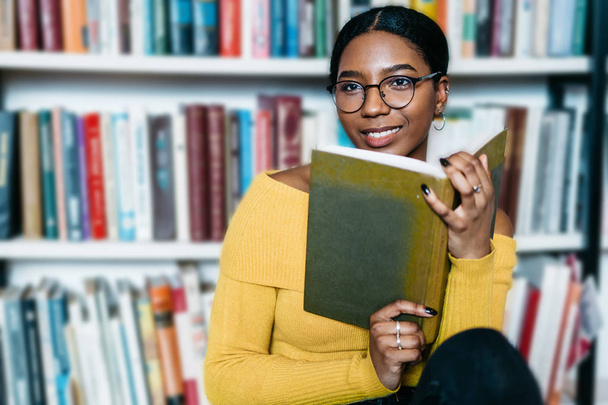 Thoughtful smiling young woman with dark skin looking away while holding book in hands.Dreamy pretty african american student thinking on literature plot of bestseller spending leisure time in library - Фото, зображення