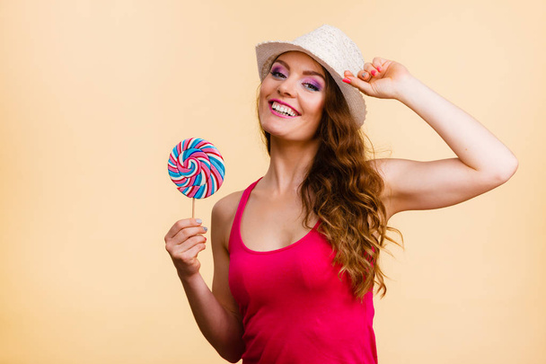 Beautiful woman wearing red tshirt summer hat holding big lollipop candy in hand. Sweet food fun concept. Studio shot on bright beige - Photo, image