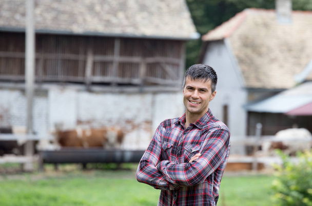 Granjero guapo seguro con brazos cruzados de pie frente a vacas y graneros simmentales en el rancho
 - Foto, imagen