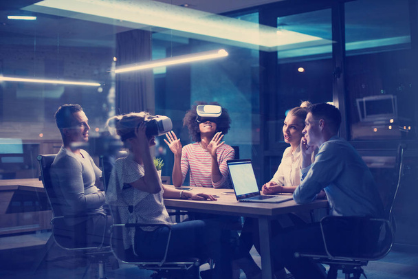 Πολυεθνική ομάδα επιχειρήσεων χρησιμοποιώντας ακουστικά εικονικής πραγματικότητας σε νυχτερινή συνάντηση γραφείου Προγραμματιστές συνάντηση με προσομοιωτή εικονικής πραγματικότητας γύρω από το τραπέζι στο δημιουργικό γραφείο. - Φωτογραφία, εικόνα