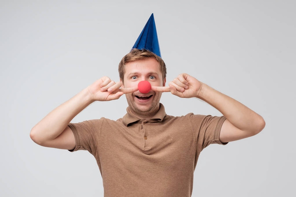 Caucasian young man with holiday cap on head and red clown nose grimacing and having fun. Concept of birthday party - Photo, Image