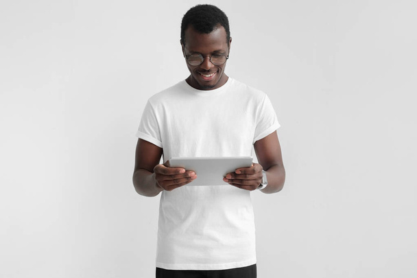 Horizontal photo of handsome African male standing isolated on gray background wearing eyeglasses and using tablet computer that he is holding, smiling, satisfied with content he sees on screen - Foto, Imagem