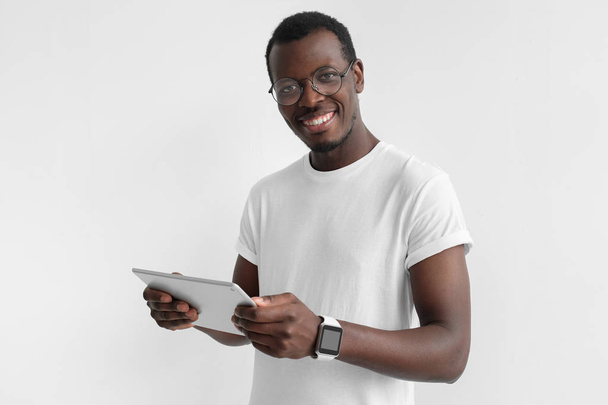 Photo rapprochée de jeune Afro-Américain photographié isolé sur fond gris debout avec tablette PC dans les mains, regardant la caméra, naviguant quelque chose sur Internet ou communiquant avec des camarades
 - Photo, image