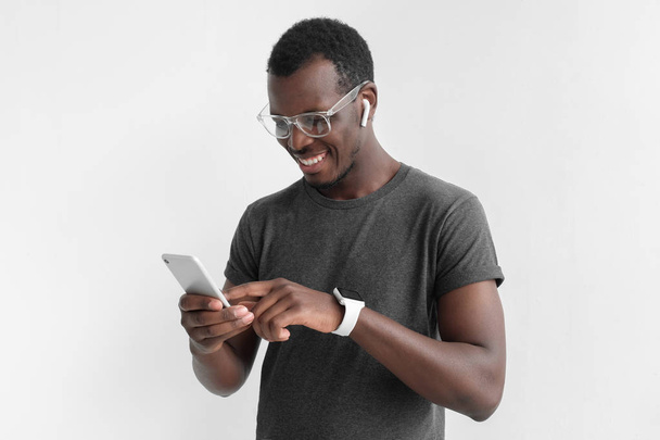 Indoor photo of young African American guy pictured isolated on grey background wearing plastic glasses and wristwatch, typing something on touchscreen of his smartphone with positive relaxed smile - Фото, зображення