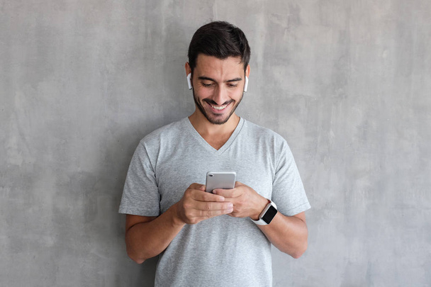 Young man wearing wireless earphones and smart watches, standing against gray textured wall, looking at screen of smartphone, browsing web and smiling nicely while chatting - Φωτογραφία, εικόνα