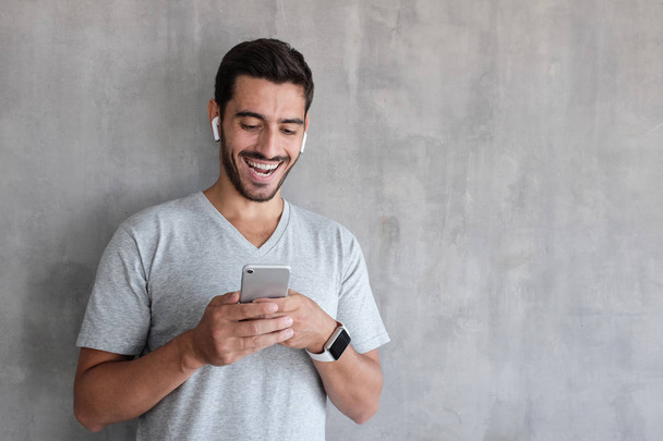 Indoor daylight picture of handsome man wearing gray casual t-shirt, laughing happily being amused by content on screen of smartphone he is holding in both hands, standing against textured wall - Photo, image