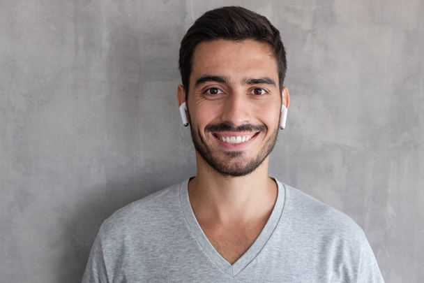 Young handsome man standing against gray textured wall, wearing t shirt and white wireless earphones, showing relaxed smile, looking confident and satisfied - Foto, Imagen