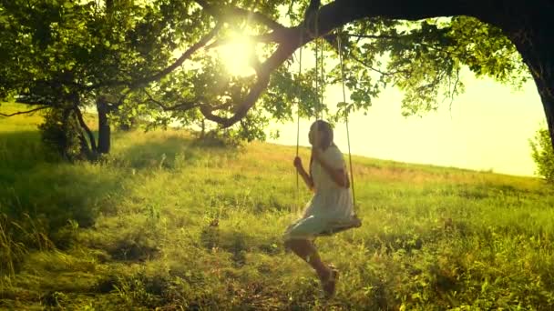 Happy girl with long hair, swinging on swing in bright rays of golden sunset and smiling. Slow motion - Footage, Video