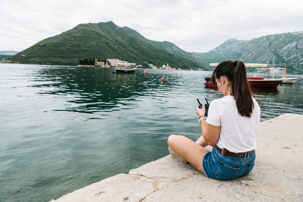 jonge volwassen vrouw zittend op pier met schitterend uitzicht op zee nd bergen. het controlemechanisme van de drone. Privaatleven - Foto, afbeelding