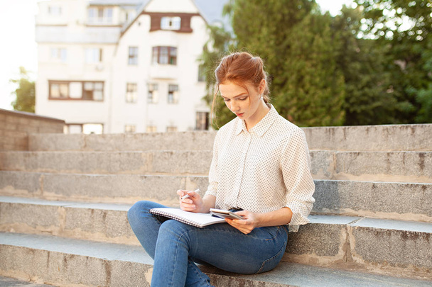 Young beautiful red hair girl with freckles sitting on a stairs near the university writing in a notebook with homework. Portrait of a student. Back to school concept. - 写真・画像