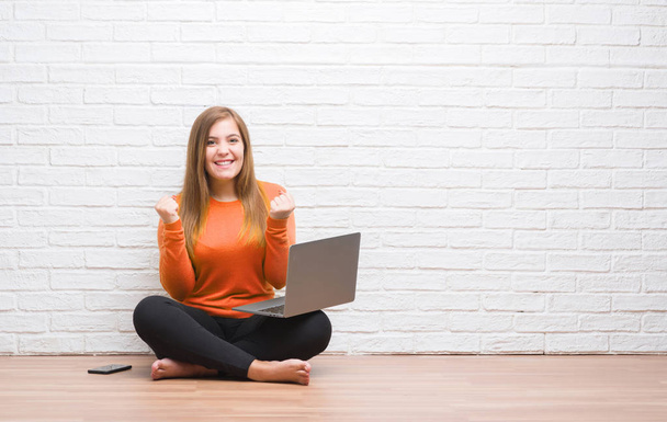 Mujer adulta joven sentada en el suelo sobre la pared de ladrillo blanco usando computadora portátil gritando orgulloso y celebrando la victoria y el éxito muy emocionado, animando la emoción
 - Foto, imagen