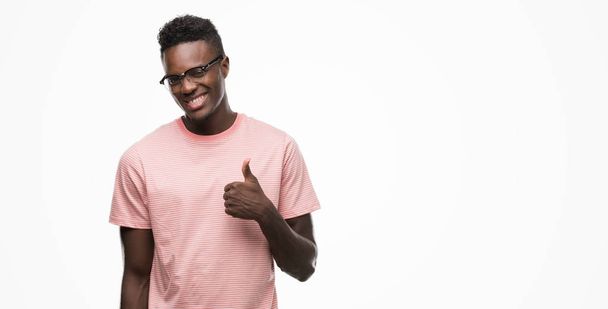 Joven hombre afroamericano vistiendo camiseta rosa haciendo felices pulgares hacia arriba gesto con mano. Aprobar la expresión mirando a la cámara con éxito
. - Foto, imagen