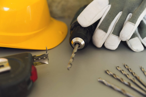 Стандарт безпеки обладнання. Жовтий твердий капелюх, рукавички, будівельна вимірювальна стрічка та електрична викрутка на сірому столі
 - Фото, зображення