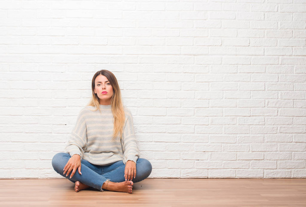 Young adult woman sitting on the floor over white brick wall at home with serious expression on face. Simple and natural looking at the camera. - Foto, imagen