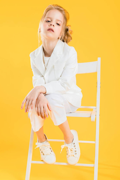 Teini-ikäinen tyttö valkoisissa vaatteissa istuu tuolilla keltaisella taustalla.
. - Valokuva, kuva