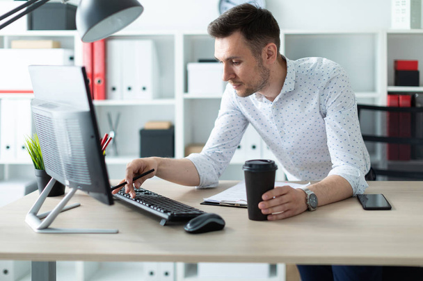 A young man is standing near a table in the office, holding a pencil and a glass of coffee. A young man works with documents and a computer. - Foto, imagen
