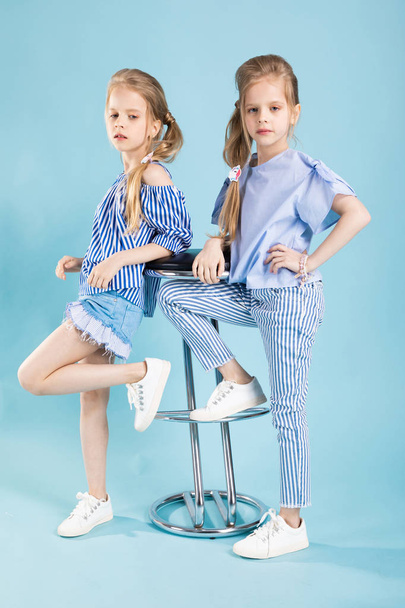 Girls twins in light blue clothes are posing near a bar stool on a blue background. - Photo, Image