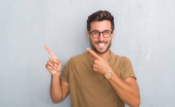 Bonito jovem sobre parede grunge cinza usando óculos sorrindo e olhando para a câmera apontando com duas mãos e dedos para o lado
. - Foto, Imagem