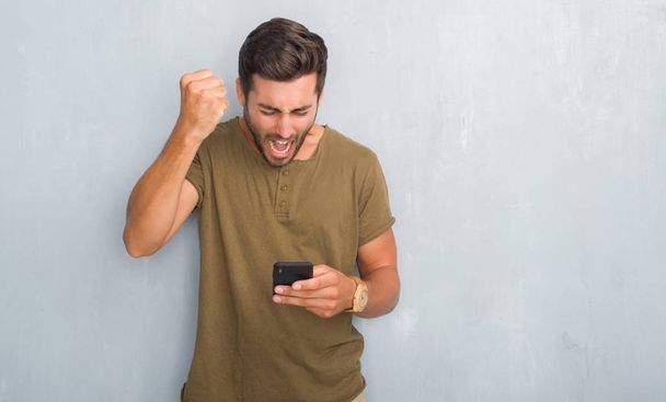Beau jeune homme sur le mur gris grunge texter un message à l'aide d'un smartphone ennuyé et frustré crier avec colère, fou et crier avec la main levée, concept de colère
 - Photo, image