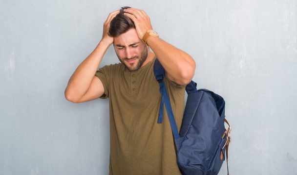 Beau touriste jeune homme sur le mur gris grunge portant sac à dos souffrant de maux de tête désespérés et stressés parce que la douleur et la migraine. Mains sur la tête
. - Photo, image