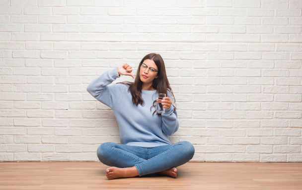 Young brunette woman sitting on the floor drinking glass of water with angry face, negative sign showing dislike with thumbs down, rejection concept - Φωτογραφία, εικόνα