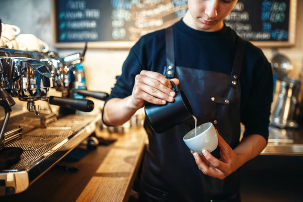 Barista in apron pours cream into the cup of coffee, cafe counter on background. Professional cappuccino preparation by bartender - Photo, Image