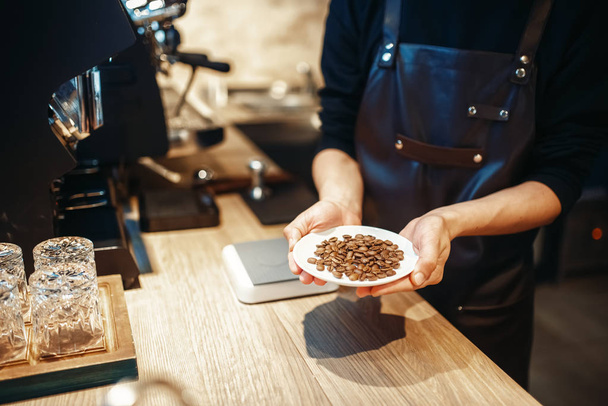 バリスタは、木製カウンターの背景に、新鮮なコーヒー豆とプレートを保持しています。カフェテリアでバーテンダー、職場でエプロンでバーテンダーによってプロのエスプレッソの準備 - 写真・画像