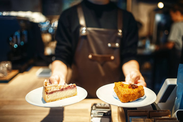 Jonge mannelijke barista holding in handen platen met zoete dessert, café teller op achtergrond. Professionele koffie voorbereiding door barman in cafetaria, barman bezetting - Foto, afbeelding