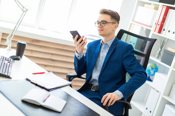 Een jonge man zit met een telefoon in zijn handen aan een tafel in het kantoor. - Foto, afbeelding
