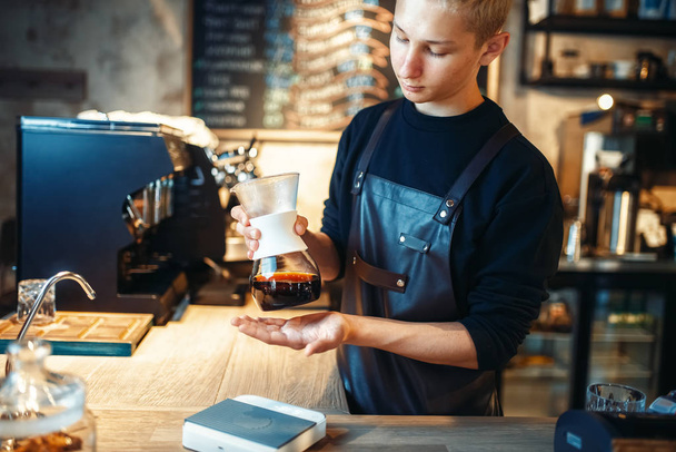 O jovem barista faz café fresco no balcão do café. Barman trabalha na cafetaria, bartender prepara café preto
 - Foto, Imagem