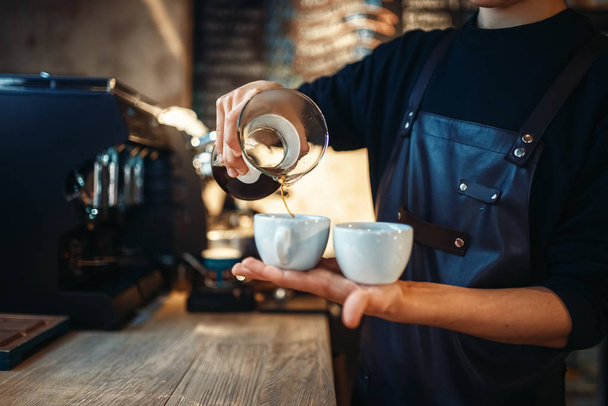 Young male barista in black apron holds in hands two cups of fresh coffee, cafe counter on background. Professional espresso preparation by barman in cafeteria, bartender occupation - Photo, Image
