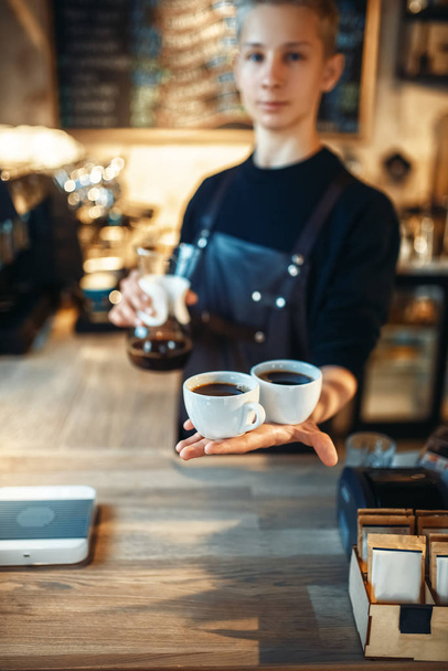 Young male barista in black apron holds in hands two cups of fresh coffee, cafe counter on background. Professional espresso preparation by barman in cafeteria, bartender occupation - Photo, Image