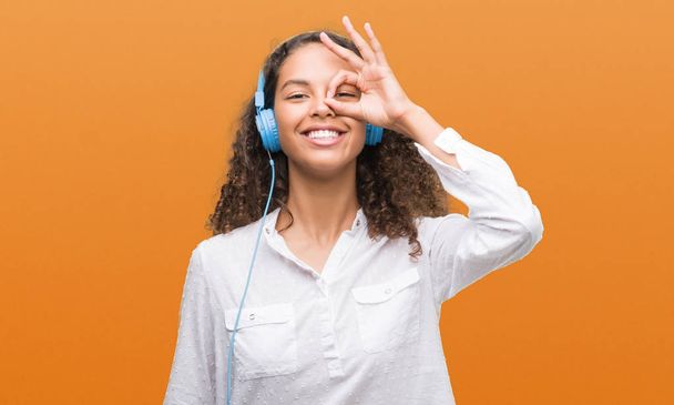 Young hispanic woman wearing headphones with happy face smiling doing ok sign with hand on eye looking through fingers - Zdjęcie, obraz
