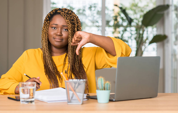Mujer afroamericana estudiando con portátil con la cara enojada, signo negativo que muestra aversión con los pulgares hacia abajo, concepto de rechazo
 - Foto, Imagen
