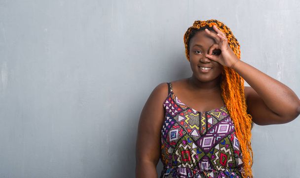 Jonge african american vrouw over grijs grunge muur dragen oranje vlechten doen ok gebaar met hand glimlachen, oog kijken door vingers met blij gezicht. - Foto, afbeelding