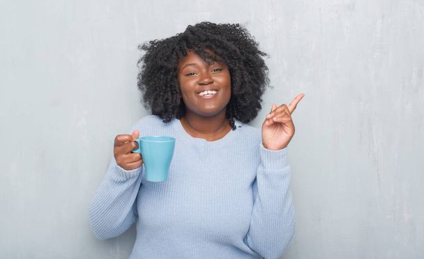 Jovem afro-americana sobre parede grunge cinza bebendo uma xícara de café muito feliz apontando com a mão e o dedo para o lado
 - Foto, Imagem