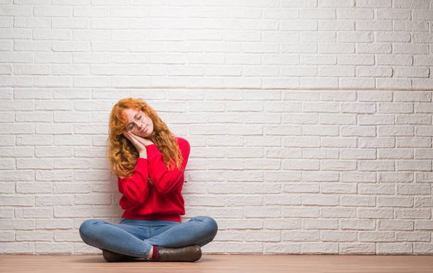 Young redhead woman sitting over brick wall sleeping tired dreaming and posing with hands together while smiling with closed eyes. - Photo, Image