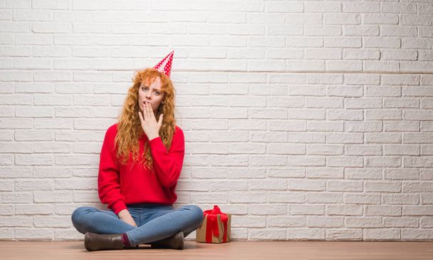 Молодая рыжая женщина сидит на кирпичной стене в шляпе на день рождения рот с рукой шокирован стыдом за ошибку, выражение страха, страшно в тишине, тайная концепция
 - Фото, изображение
