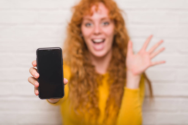 Jeune femme rousse assise sur un mur de briques montrant smartphone très heureux et excité, expression gagnante célébrant la victoire en criant avec un grand sourire et les mains levées
 - Photo, image