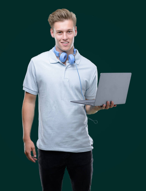 Young handsome blond man using a laptop with a happy face standing and smiling with a confident smile showing teeth - Photo, image