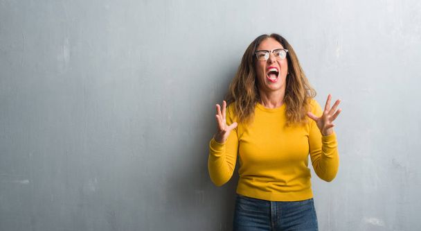 Mujer hispana de mediana edad sobre una pared gris con gafas locas y locas gritando y gritando con expresión agresiva y brazos levantados. Concepto de frustración
. - Foto, imagen