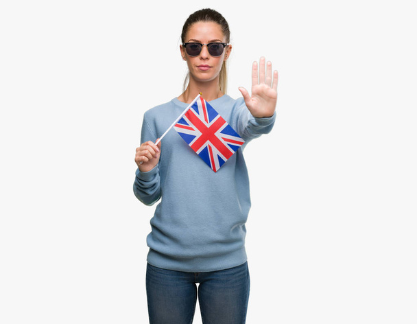 Mulher bonita segurando bandeira do Reino Unido com a mão aberta fazendo sinal de parada com expressão séria e confiante, gesto de defesa
 - Foto, Imagem