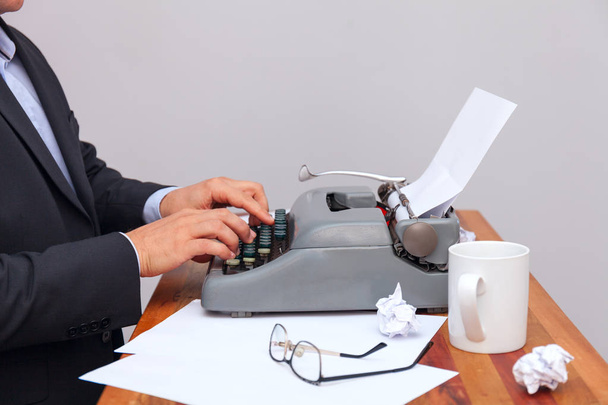 Der Mann im Anzug tippt auf einer altmodischen Schreibmaschine. Auf dem Tisch stehen Gläser und ein Becher. neutraler Hintergrund - Foto, Bild