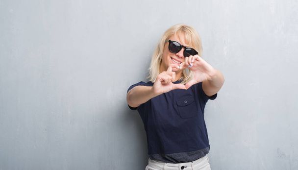 Adult caucasian woman over grunge grey wall wearing sunglasses smiling in love showing heart symbol and shape with hands. Romantic concept. - Fotó, kép