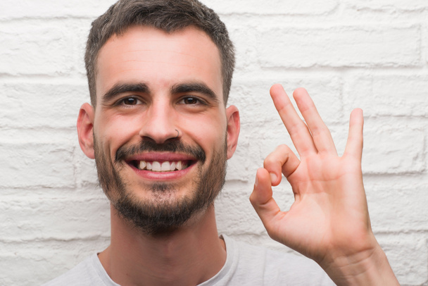 Genç yetişkin adam parmak, mükemmel sembolü ile ok işareti yapıyor beyaz tuğla duvarın üzerinden ayakta - Fotoğraf, Görsel