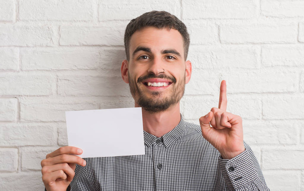 Homem adulto jovem sobre parede de tijolo segurando papel em branco surpreso com uma ideia ou pergunta apontando dedo com rosto feliz, número um
 - Foto, Imagem