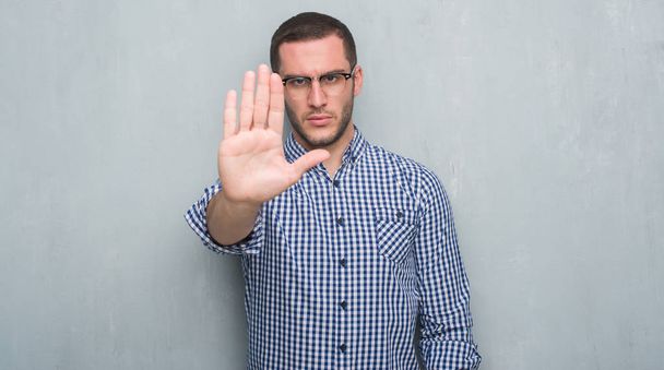 Young caucasian business man over grey grunge wall with open hand doing stop sign with serious and confident expression, defense gesture - Photo, Image