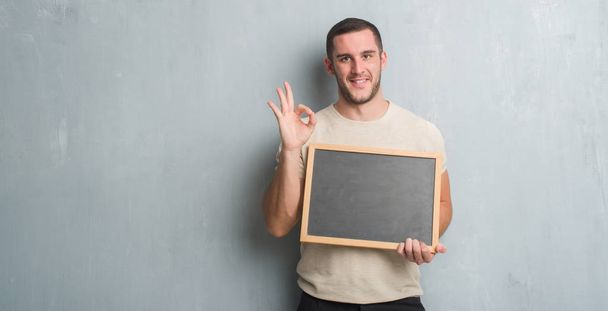 Kaukasische jongeman over grijs grunge muur holding schoolbord doen ok teken met vingers, uitstekende symbool - Foto, afbeelding